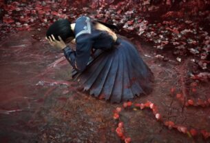 Hinako holds her head in her hand as she lays on the ground surrounded by red plants.
